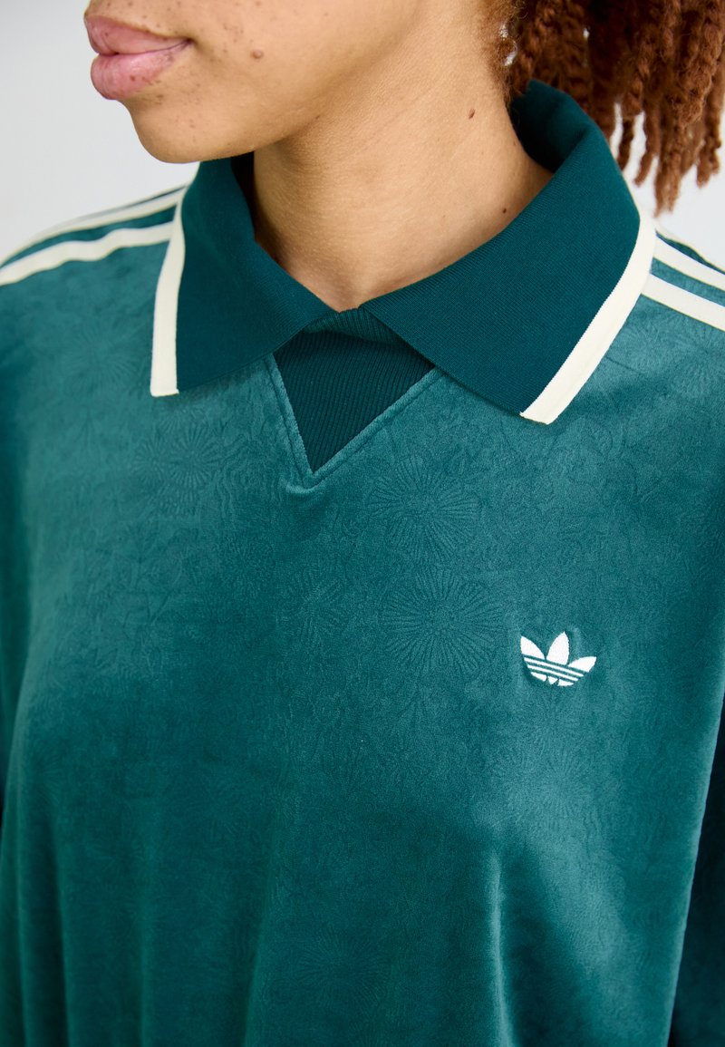 Persoon met een groene fluwelen Adidas-shirt met een wit logo, groene kraag en crème strepen op de schouder, weergegeven van borsthoogte naar boven.