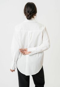 Fehér, hosszú ujjú ing, texturált anyaggal, hátrahúzott aljjal és gombos mandzsettával. Hátul ráncolt rész található, amely mozgékonyságot biztosít.