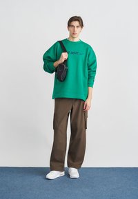 Πράσινο φούτερ με στρογγυλή λαιμόκοψη και το κείμενο «JUST DO IT» σε μπλε γράμματα, καφέ παντελόνι καρότσα, λευκά αθλητικά παπούτσια και μια μικρή μαύρη τσάντα χιαστί.