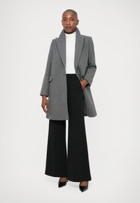 Palton gri din lână cu guler tip notch și buzunare laterale, asortat cu un pulover alb cu guler înalt și pantaloni negri evazați. Pantofii cu toc negri adaugă un contrast.