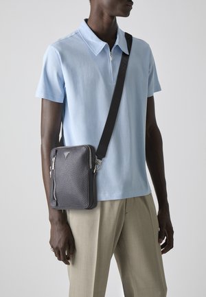 Grau strukturierte Umhängetasche mit silbernen Reißverschlüssen, verstellbarem Trageriemen und dreieckigem Logo; getragen über einem hellblauen Poloshirt und beigen Hosen.