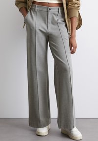 Pantalons larges gris avec des plis, fabriqués en tissu doux. Présente des poches latérales et une fermeture à bouton à l'avant. Associés à des baskets blanches.