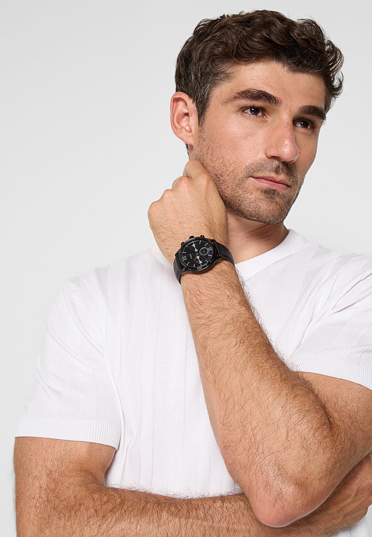 Reloj analógico negro con dial texturizado y acentos plateados en contraste, llevado en una muñeca, frente a un fondo de una camisa blanca de manga corta con rayas.