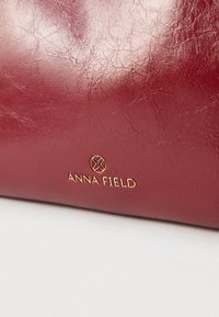 Sac à main en similicuir rouge avec une surface texturée et un logo doré "ANNA FIELD". Design compact avec des bords arrondis.