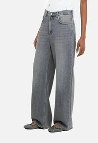 Wide-leg, high-waisted jeans in gray denim with front pockets and subtle stitching details. Model paired with black embellished flats.
