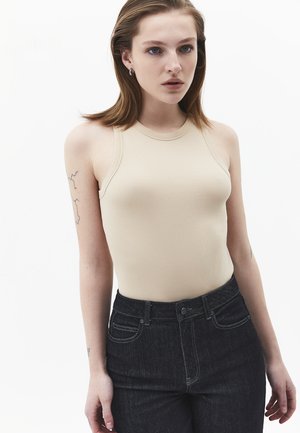 Giovane donna con capelli castano chiaro che indossa un top beige senza maniche a coste e jeans scuri a vita alta, in piedi davanti a uno sfondo bianco uniforme.