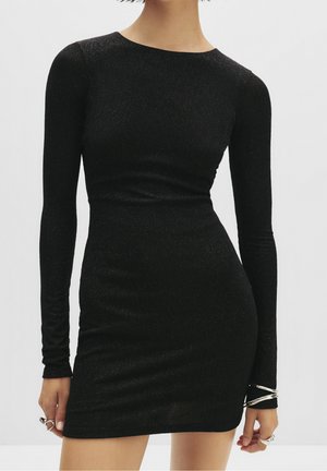 Femme portant une mini-robe noire cintrée à manches longues avec un léger scintillement, accessoirisée de bagues et bracelets argentés aux deux mains.