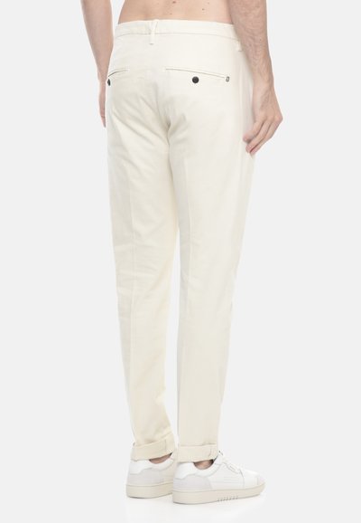Uomo con pantaloni tagliati color avorio con tasche posteriori con bottone e sneakers bianche, in piedi contro uno sfondo neutro.