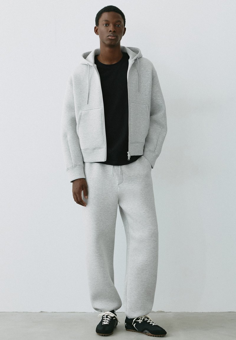 Jeune homme debout contre un mur blanc, portant un sweat à capuche gris clair et un pantalon de survêtement avec un t-shirt noir et des baskets noires aux lacets blancs.