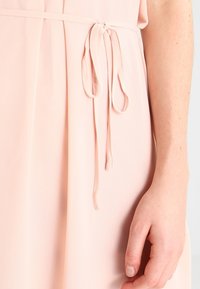 Robe rose pâle avec une texture douce et fluide. Une ceinture nouée, réalisée dans le même matériau, est mise en avant.