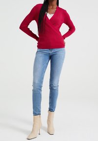 Červený žebrovaný zavinovací svetr s dlouhými rukávy přes krajkový top na ramínka, v kombinaci se světle modrými úzkými džíny a béžovými kotníkovými botami na podpatku.