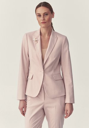 Kvinde med tilbagetrukket hår iført en sart lyserød skræddersyet dragt og blomsterbroche, stående mod en ensfarvet lys baggrund.