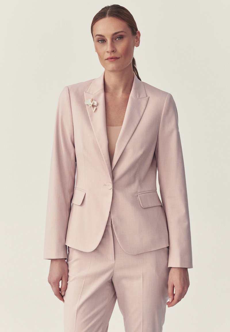 Femme aux cheveux tirés en arrière, portant un tailleur rose pâle ajusté et une broche florale, debout devant un fond clair uni.