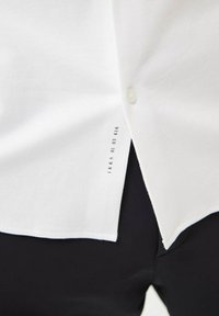 Witte katoenen shirt met knoopsluiting, afgeronde onderkant en bedrukte label aan de zijkant, gecombineerd met zwarte broeken.