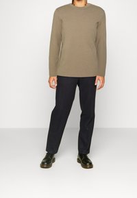 Maglione beige a maniche lunghe con scollo rotondo abbinato a pantaloni blu scuro e scarpe nero stringate, in piedi contro uno sfondo bianco semplice.