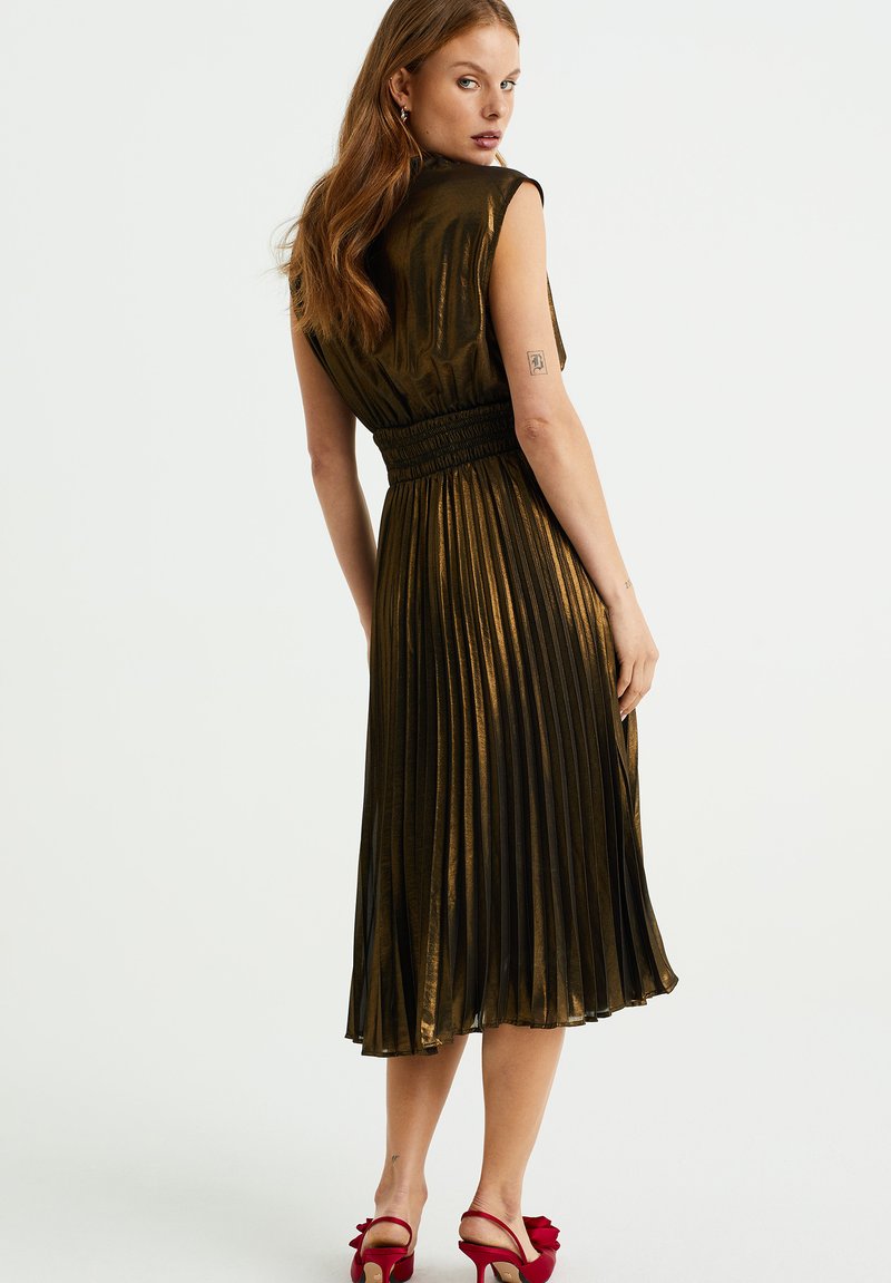 Donkergroene geplooide jurk met een getailleerde taille, korte mouwen en een glanzende textuur, gecombineerd met rode haksandalen.