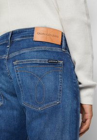 Blaue Denim-Jeans mit einem hellbraunen Lederbundetikett, hinteren Aufsetztaschen mit kontrastierendem Stich und einem markanten Schlaufendesign an der Tasche.