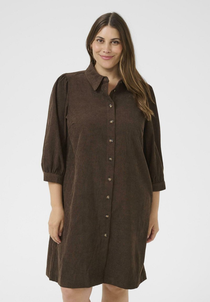 Lächelnde Frau in einem braunen Knopfkleid mit Kragen und dreiviertel Puffärmeln, die vor einem schlichten hellen Hintergrund steht.