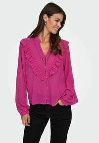 Fuchsia Bluse mit Bausch vorne, Druckknopfverschluss und langen Ärmeln, kombiniert mit schwarzen Hosen. Weich, texturiert.