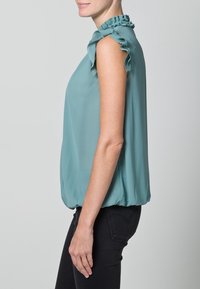 Top senza maniche di colore teal con dettagli arricciati sulle spalle, texture morbida e vestibilità ampia, raccolto all'orlo, indossato con pantaloni neri.
