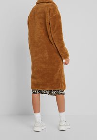 Brun, fluffy, mellemlang frakke med høj krave. Bæres over en leopardmønstret kjole, parret med hvide sokker og sneakers.