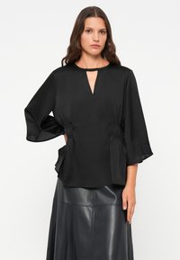 Zwarte blouse met een keyhole-halslijn, brede uitlopende mouwen en geplooide zijdetails. De stof lijkt glad met een lichtgewicht textuur.