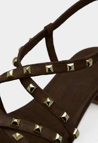 Καφέ σουέτ σανδάλι με διασταυρωμένα λουράκια, διακοσμημένο με μεταλλικά χρυσά πυραμιδοειδή καρφιά κατά μήκος των λουριών και με υφή στο πέλμα.