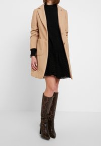 Camel-farbener Wollmantel mit zwei Vorderaschen, über einem schwarzen strukturierten Kleid getragen, kombiniert mit kniehohen Stiefeln im Schlangenprint.