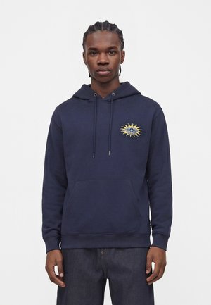 Jeune homme aux cheveux tressés portant un sweat à capuche bleu marine avec un logo jaune de soleil et d'œil sur la poitrine, debout devant un fond uni.