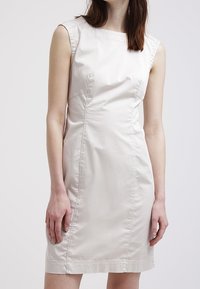 Över knäet, ärmlös klänning i ljus beige tyg. Har en rund halsringning, figursydd design och subtil sömdetaljering genom hela klänningen.