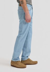 Hombre con jeans azul claro de pierna recta y zapatos casuales de ante color beige, de pie contra un fondo liso.