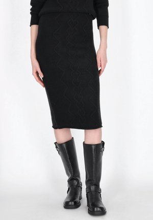 Personne portant une jupe noire texturée longueur genou et des bottes en cuir noires montantes avec des boucles, debout devant un fond blanc.
