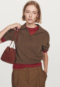 Ung kvinde med kort blond hår iført lagdelte brune og røde toppe samt brune bukser, bærende en rødbrun suede håndtaske over skulderen.