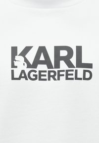Bílá mikina s výrazným černým nápisem "KARL LAGERFELD" a grafickým siluetou postavy nosící sluneční brýle.