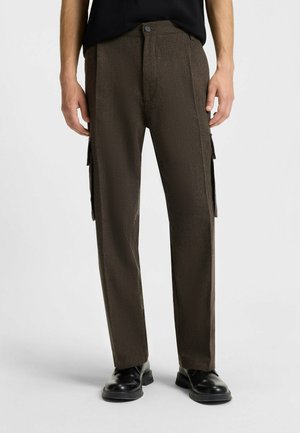 Cargo-Hosen aus dunkelbraunem, strukturiertem Stoff mit geradem Schnitt, ausgestattet mit Seitentaschen und einer vorderen Knopfleiste. Schwarze Schuhe runden den Look ab.