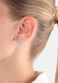 Primo piano dell'orecchio di una donna bionda con un piccolo orecchino a cerchio in argento texture, capelli raccolti all'indietro, che indossa una camicia bianca con colletto.