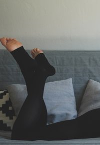 Black opaque leggings on a light grey sofa, showing bare feet with a rounded shape. Two grey pillows are in the background, one patterned.