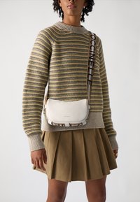 Bílá kožená crossbody taška s hladkou texturou a širokým popruhem, na němž je vytištěn název značky tučnými černými písmeny. Nošena s pruhovaným svetrem a sukní.