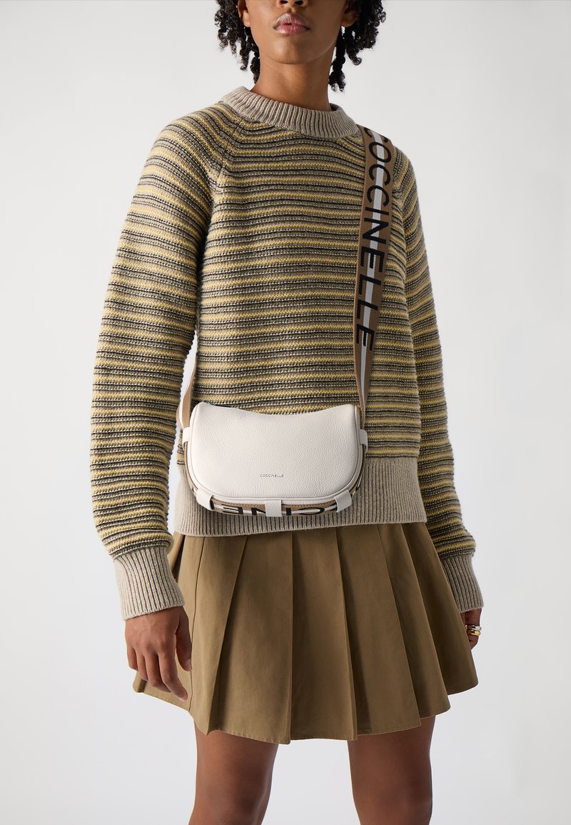 Bílá kožená crossbody taška s hladkou texturou a širokým popruhem, na němž je vytištěn název značky tučnými černými písmeny. Nošena s pruhovaným svetrem a sukní.