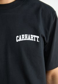 Czarna bawełniana koszulka z białym, zakrzywionym logo "CARHARTT" nadrukowanym na piersi. Krótkie rękawy i klasyczny dekolt typu crew neck.