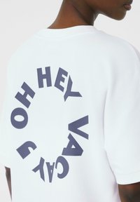 Biała koszulka z okrągłym dekoltem z fioletowym graficznym napisem ułożonym w okrągły wzór na plecach. Materiał bawełniany, krótki rękaw.
