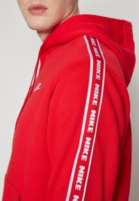 Rdeča kapuca z velikim sprednjim žepom, kapuco z vrvico in belim logotipom Nike ter črtasto poudarjeno oblogo ob rami.