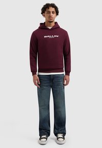 Bordeauxrode hoodie met "BALLIN'" logo, gecombineerd met losse blauwe spijkerbroek en zwart-witte sneakers. Casual pasvorm en gestructureerde stof.