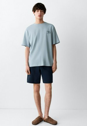 Jonge man staand, gekleed in een lichtblauw oversized T-shirt met de tekst "Always Fresh Served Daily", marineblauwe korte broek en bruine instapschoenen.