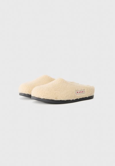 Beige tofflor i päls med rund tå, svart gummisula och en liten röd och vit etikett som visar varumärket.