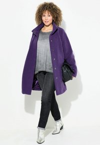 Abrigo de forro polar morado con cuello alto y cierre de botones, llevado sobre una blusa gris y jeans oscuros. Combinado con botas de tobillo plateadas brillantes.