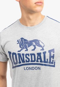 Pilkas trumpas rankovėmis marškinėlis, pagamintas iš medvilnės, su mėlynu liūto grafika ir ryškiu tekstu "LONSDALE LONDON" priekyje.