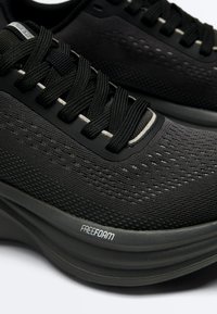 Gros plan sur des chaussures de course en maille noire avec des lacets noirs et l'inscription "FREEFOAM" sur la semelle épaisse et amortissante.