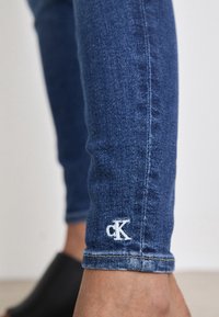 Jeans de denim azul ajustados con costuras claras y un pequeño logo blanco "ck" en el tobillo. Textura suave y diseño ceñido.