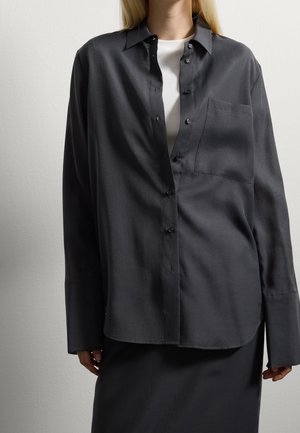Femme portant une chemise ample et sombre à boutons sur un haut blanc, associée à une jupe foncée, debout devant un mur clair uni.